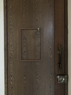 A wooden door with a prominent wood grain texture features a small square hatch in the center. The latch of the hatch is visible, designed for securing it shut. On the right side of the door, a vintage-style metal lock is installed along with an intricate handle.
