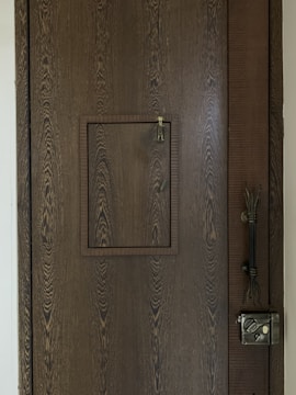 A wooden door with a prominent wood grain texture features a small square hatch in the center. The latch of the hatch is visible, designed for securing it shut. On the right side of the door, a vintage-style metal lock is installed along with an intricate handle.
