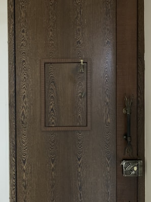 A wooden door with a prominent wood grain texture features a small square hatch in the center. The latch of the hatch is visible, designed for securing it shut. On the right side of the door, a vintage-style metal lock is installed along with an intricate handle.