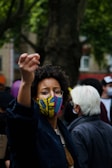 A person wearing a colorful facemask raises their hand in a gesture, suggesting engagement or protest. Surrounding them are other people, one with white hair, possibly at a public gathering or demonstration. Trees and blurred urban background suggest an outdoor city setting.