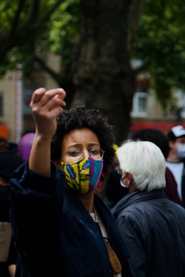 A person wearing a colorful facemask raises their hand in a gesture, suggesting engagement or protest. Surrounding them are other people, one with white hair, possibly at a public gathering or demonstration. Trees and blurred urban background suggest an outdoor city setting.