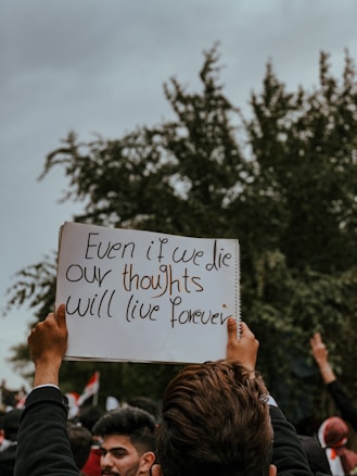 A person holds up a handwritten sign that reads 'Even if we die our thoughts will live forever' amidst a crowd. The background features trees and a cloudy sky, with other people and flags partially visible.