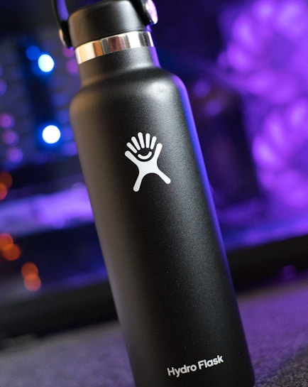 A black stainless steel Hydro Flask water bottle with a silver cap and a white logo featuring a stylized hand with a wave. The background is blurred with bokeh lighting, predominantly purple, adding a soft, ambient feel.