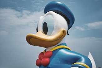 A large inflatable duck character wearing a blue sailor suit and hat, with a red bow tie, set against a partly cloudy sky.