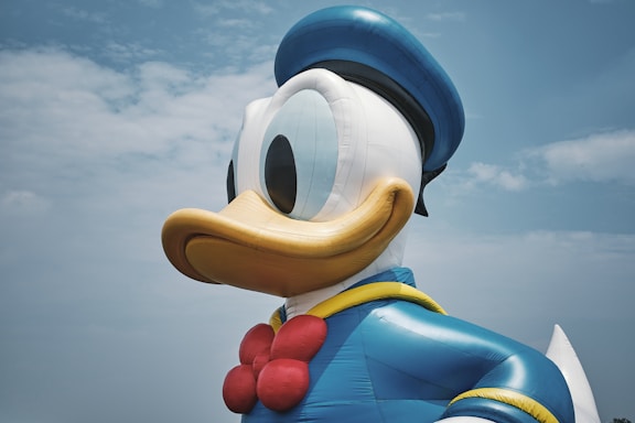 A large inflatable duck character wearing a blue sailor suit and hat, with a red bow tie, set against a partly cloudy sky.