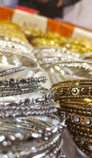 A close-up of intricately designed metal bangles featuring silver and gold hues, adorned with small embedded stones. The focus on the delicate details highlights their ornate craftsmanship. The background is slightly blurred, emphasizing the foreground bangles.