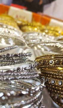 A close-up of intricately designed metal bangles featuring silver and gold hues, adorned with small embedded stones. The focus on the delicate details highlights their ornate craftsmanship. The background is slightly blurred, emphasizing the foreground bangles.