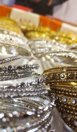 A close-up of intricately designed metal bangles featuring silver and gold hues, adorned with small embedded stones. The focus on the delicate details highlights their ornate craftsmanship. The background is slightly blurred, emphasizing the foreground bangles.