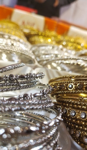 A close-up of intricately designed metal bangles featuring silver and gold hues, adorned with small embedded stones. The focus on the delicate details highlights their ornate craftsmanship. The background is slightly blurred, emphasizing the foreground bangles.