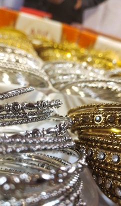 A close-up of intricately designed metal bangles featuring silver and gold hues, adorned with small embedded stones. The focus on the delicate details highlights their ornate craftsmanship. The background is slightly blurred, emphasizing the foreground bangles.