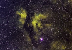 A vast field of stars scattered across the cosmos with swirling clouds of gas and dust in hues of green and yellow, likely representing a nebula. The cosmic dust creates patterns against the dark backdrop, with some bright star clusters and individual stars prominently visible.