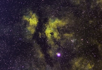 A vast field of stars scattered across the cosmos with swirling clouds of gas and dust in hues of green and yellow, likely representing a nebula. The cosmic dust creates patterns against the dark backdrop, with some bright star clusters and individual stars prominently visible.
