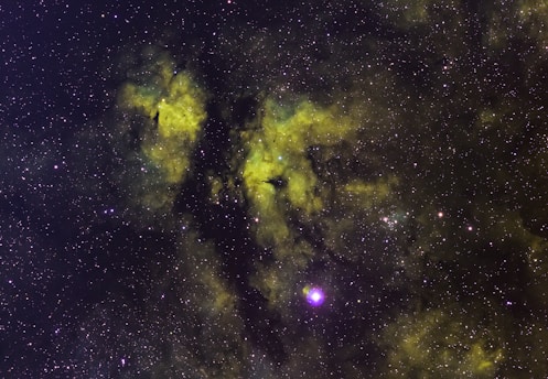 A vast field of stars scattered across the cosmos with swirling clouds of gas and dust in hues of green and yellow, likely representing a nebula. The cosmic dust creates patterns against the dark backdrop, with some bright star clusters and individual stars prominently visible.