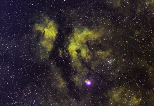 A vast field of stars scattered across the cosmos with swirling clouds of gas and dust in hues of green and yellow, likely representing a nebula. The cosmic dust creates patterns against the dark backdrop, with some bright star clusters and individual stars prominently visible.