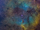 A vibrant and colorful section of a nebula filled with countless stars against a cosmic background. Intricate dark patterns weave through shades of teal, gold, and green, creating a dynamic and lively cosmic scene with varying textures and densities.