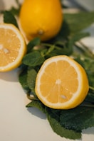 Two yellow lemons are placed on top of fresh green mint leaves. One lemon is whole while the other is cut in half, revealing its juicy interior and seeds.