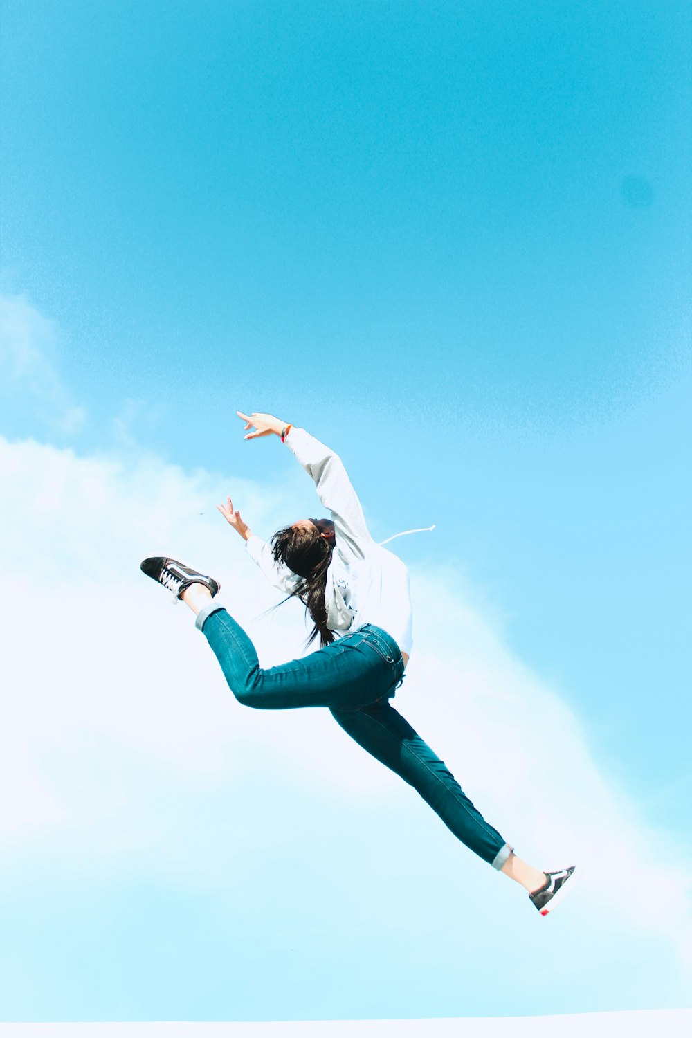 Mujer con camisa blanca de manga larga y jeans de mezclilla azul saltando bajo el cielo azul durante el día