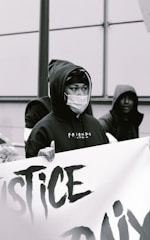 A person wearing a hooded sweatshirt and a face mask is holding a large banner with partially visible text. The scene appears to be a protest or demonstration, with a focus on justice-related themes. Other individuals are present, also wearing hoodies.