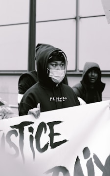 A person wearing a hooded sweatshirt and a face mask is holding a large banner with partially visible text. The scene appears to be a protest or demonstration, with a focus on justice-related themes. Other individuals are present, also wearing hoodies.