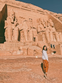 A woman stands in front of the massive stone statues and carvings of Abu Simbel, an ancient Egyptian temple. The sculptures are intricately detailed, showcasing large seated figures, likely depicting pharaohs, against the backdrop of a desert landscape. The woman is smiling, wearing casual clothing with a white top and brown pants. The sky is clear and blue.