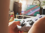 A person is holding a retro-styled video game controller with their thumbs resting on the analog sticks. In the background, a television display shows a video game scene featuring a white duck walking along a stream bordered by grass. The room is warmly lit, with various items such as a clock and some plants visible in the background.