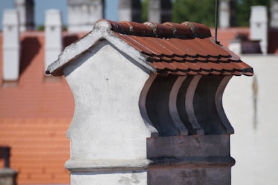 A close-up view of a chimney with a red tiled roof against a backdrop of similar architectural structures. The chimney is made of light-colored bricks, and the tiles are neatly aligned. The background is slightly blurred, highlighting the focus on the chimney.