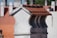 A close-up view of a chimney with a red tiled roof against a backdrop of similar architectural structures. The chimney is made of light-colored bricks, and the tiles are neatly aligned. The background is slightly blurred, highlighting the focus on the chimney.