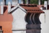 A close-up view of a chimney with a red tiled roof against a backdrop of similar architectural structures. The chimney is made of light-colored bricks, and the tiles are neatly aligned. The background is slightly blurred, highlighting the focus on the chimney.