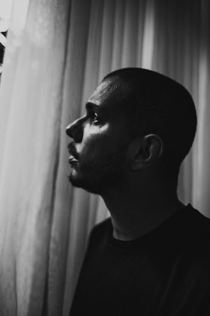 A grayscale portrait depicts a person in profile with a contemplative expression, gazing outside through sheer curtains. The lighting creates a dramatic contrast, emphasizing the contours of the face.