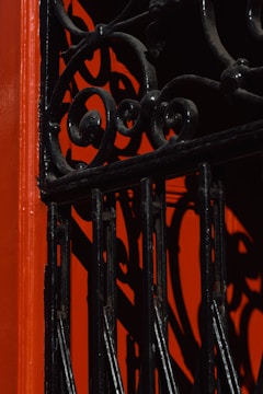 An ornamental wrought iron gate with intricate swirl patterns casts bold shadows against a vibrant red background, creating an artistic interplay of light and texture.