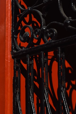 An ornamental wrought iron gate with intricate swirl patterns casts bold shadows against a vibrant red background, creating an artistic interplay of light and texture.
