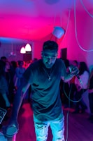 A person holding a microphone stands in the foreground of a room lit with vibrant pink and purple lights. The background shows a crowd of people, suggesting a party or music event atmosphere. The person appears engaged with the audience, adding to the lively and energetic mood.