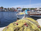 A picturesque coastal scene features fishing boats along a calm waterfront with vibrant blue water reflecting the sky. The foreground is dominated by a tangle of yellow fishing nets with red buoys. In the background, a row of quaint buildings and palm trees lines the shore under a clear blue sky, suggesting a serene and lively fishing village.