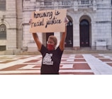A person wearing a mask and glasses holds up a cardboard sign that reads 'housing is racial justice.' The individual is standing in front of a large building with arched doorways and is wearing a shirt with text supporting Black women.