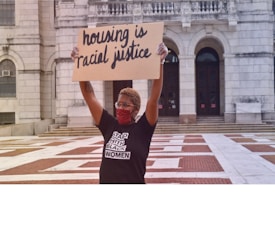 A person wearing a mask and glasses holds up a cardboard sign that reads 'housing is racial justice.' The individual is standing in front of a large building with arched doorways and is wearing a shirt with text supporting Black women.