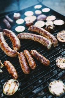 Grilled sausages and slices of vegetables such as zucchini and eggplant are cooking on a black grill. The food is cooking over an open flame with slight charring visible on the sausages and vegetables.