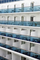 A series of stacked balconies on a modern cruise ship. Each balcony is equipped with blue-tinted glass railings and has matching white chairs and small tables. The balconies are evenly spaced and uniform, with sliding doors leading into the cabins.
