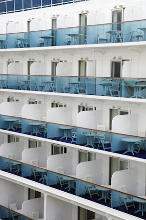 A series of stacked balconies on a modern cruise ship. Each balcony is equipped with blue-tinted glass railings and has matching white chairs and small tables. The balconies are evenly spaced and uniform, with sliding doors leading into the cabins.