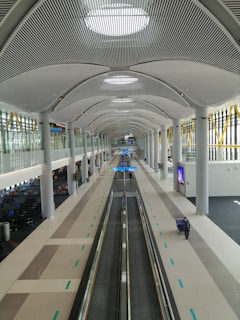A spacious and modern airport terminal with high ceilings and a long moving walkway. The architectural design features sleek lines and abundant natural light from large windows. Seating areas on the sides and a few travelers are visible.