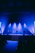 A stage is illuminated with blue lighting, featuring a band setup with musicians. The area includes instruments such as drums and microphones. The space appears to be a large auditorium with dim seating in the foreground.
