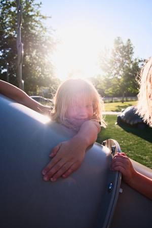 A young child with blonde hair is joyfully playing on playground equipment during a sunny day. The sunlight creates a warm backlight, highlighting the child's hair and casting a gentle glow on the scene. The background features lush green trees and a grassy area, suggesting a park setting.