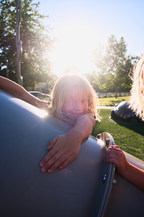 A young child with blonde hair is joyfully playing on playground equipment during a sunny day. The sunlight creates a warm backlight, highlighting the child's hair and casting a gentle glow on the scene. The background features lush green trees and a grassy area, suggesting a park setting.