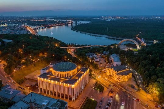An aerial view of a city at dusk, showcasing a large circular building with illuminated columns, surrounded by lush greenery and roads with a gentle traffic flow. In the background, a river snakes through the landscape, bordered by bridges and distant city lights, creating a serene and vibrant urban scene.