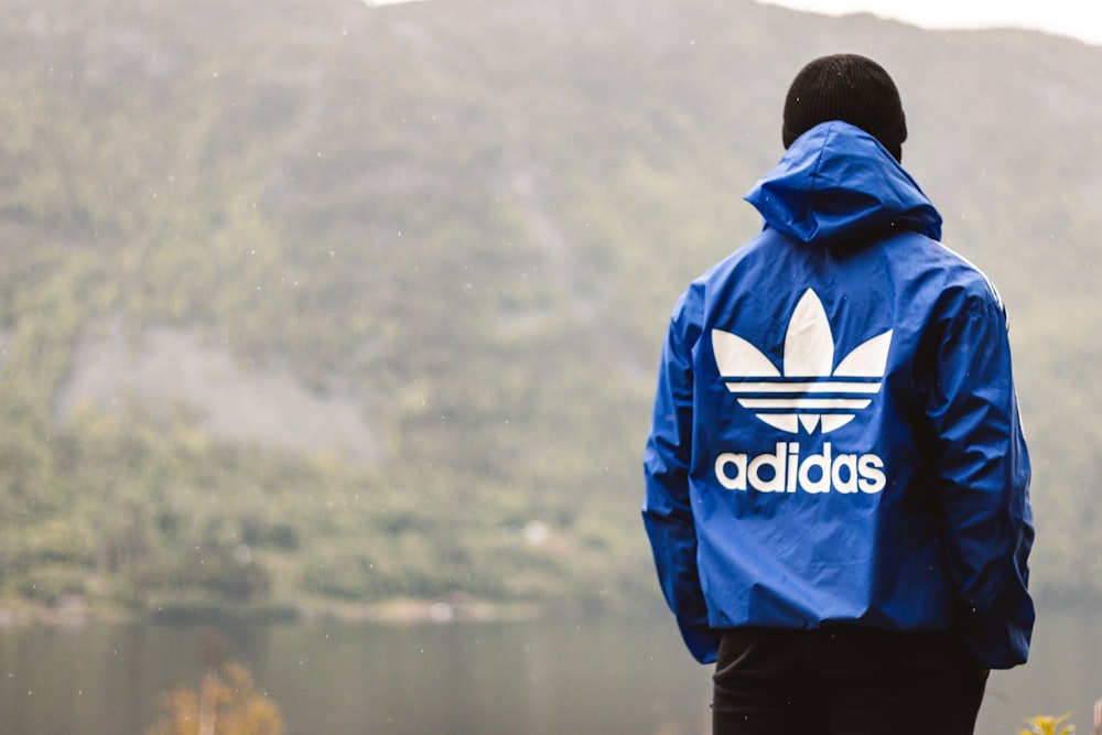 man in blue and black adidas hoodie standing near lake during daytime