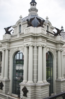 The image depicts an ornate architectural structure with large columns and arched windows. The building features a dome-like roof adorned with sculpted eagle statues at each corner. The design is reminiscent of classical architecture, with intricate detailing and a symmetrical facade.