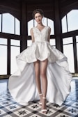 A woman stands confidently wearing a detailed white dress with a high-low hem, featuring intricate floral designs. Her hair is elegantly styled and she is wearing white high heels. The setting includes large, arched windows with a checkered floor pattern, creating a sophisticated backdrop.