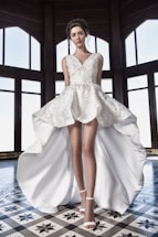 A woman stands confidently wearing a detailed white dress with a high-low hem, featuring intricate floral designs. Her hair is elegantly styled and she is wearing white high heels. The setting includes large, arched windows with a checkered floor pattern, creating a sophisticated backdrop.