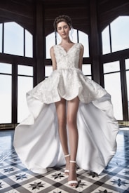 A woman stands confidently wearing a detailed white dress with a high-low hem, featuring intricate floral designs. Her hair is elegantly styled and she is wearing white high heels. The setting includes large, arched windows with a checkered floor pattern, creating a sophisticated backdrop.