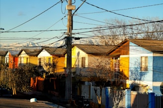 A row of colorful houses with vibrant blue, yellow, and orange facades is bathed in sunlight. Power lines crisscross the scene above, attached to a central utility pole. The houses feature gabled roofs and small front yards with trees and shrubs. In the background, snow-capped mountains are visible under a clear blue sky.