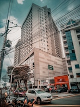 A busy urban street scene with a tall office building in the background. The sky is overcast, and power lines crisscross the upper part of the image. Several cars and motorcycles are at the foreground intersection, and there are street signs pointing towards Bogota and Cali. Trees are present along the sidewalk, and various small shops are visible at street level.